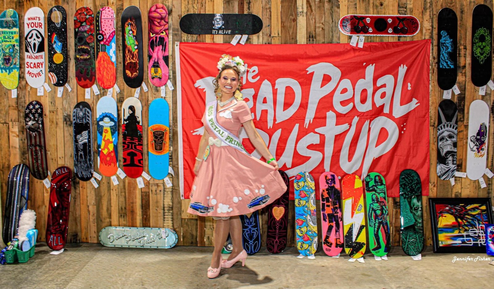 A contestant in a pink dress poses in front of The Dead Pedal Dust Up banner and a wall of painted skateboard decks